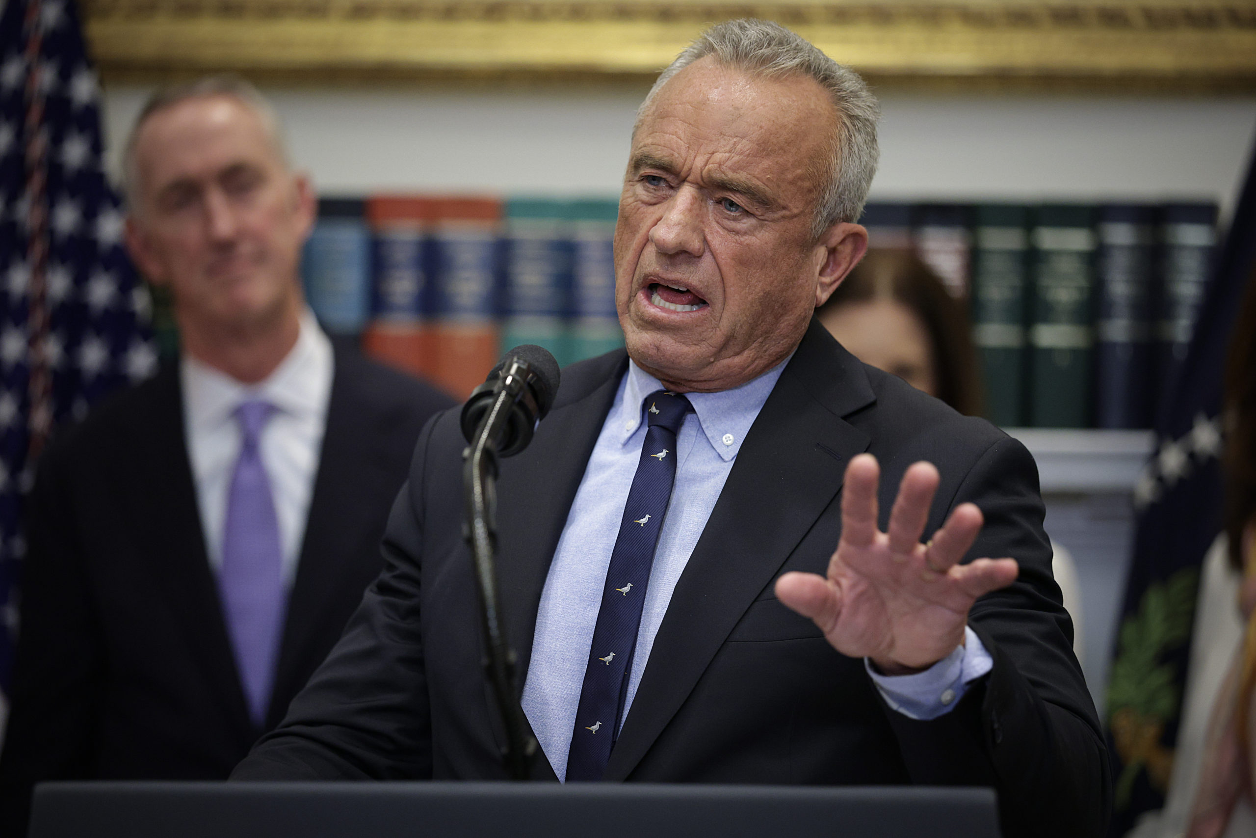 Health Secretary Robert F. Kennedy Jr., wearing a gray suit with a narrow tie, gestures while speaking into a microphone.
