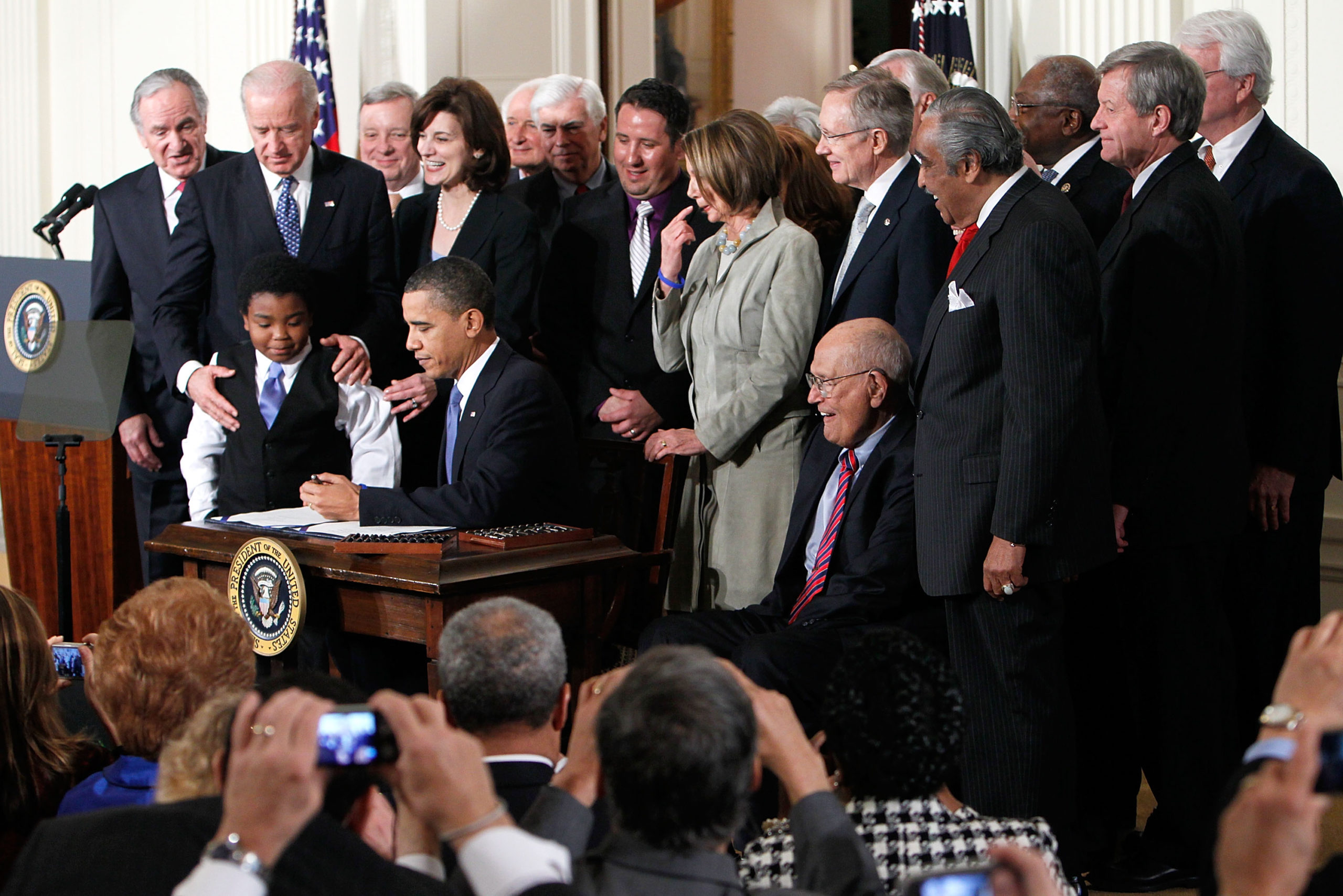 President Barack Obama sits at a desk, surrounded by politicians and aides, signing documents.