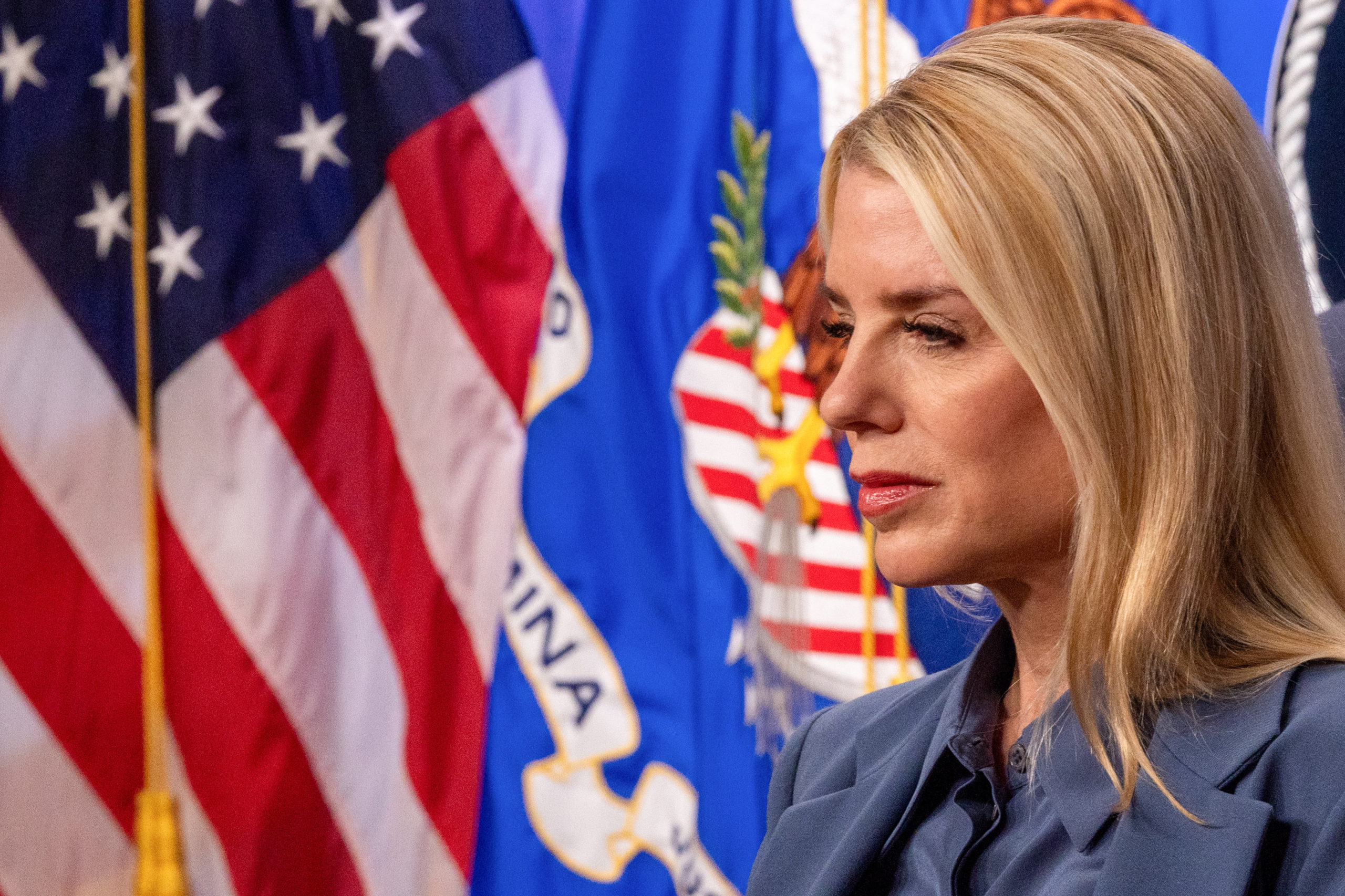 Pam Bondi, a white woman with blonde hair, is seen in profile in front of an American flag.
