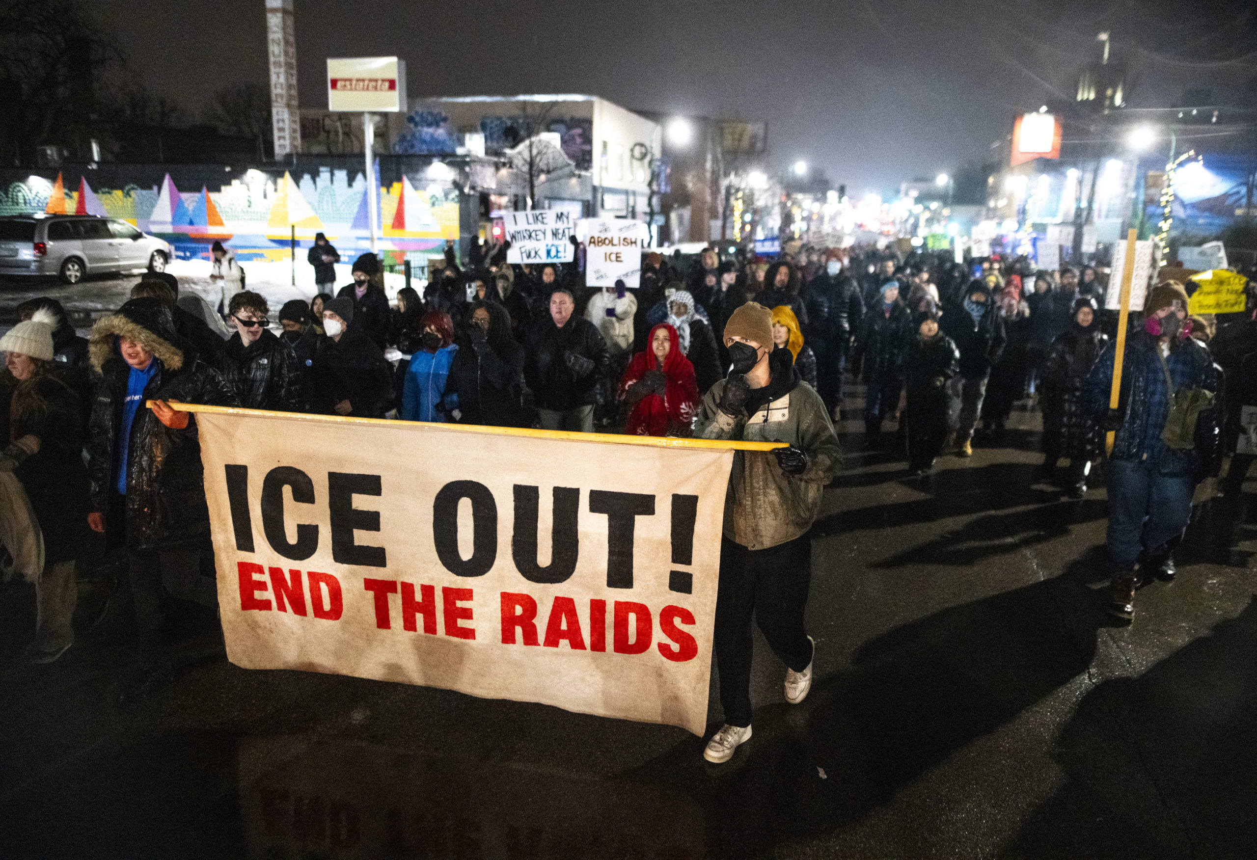 A nighttime protest in the streets of Minneapolis, some demonstrators hold a sign reading “ICE OUT! END THE RAIDS”. 