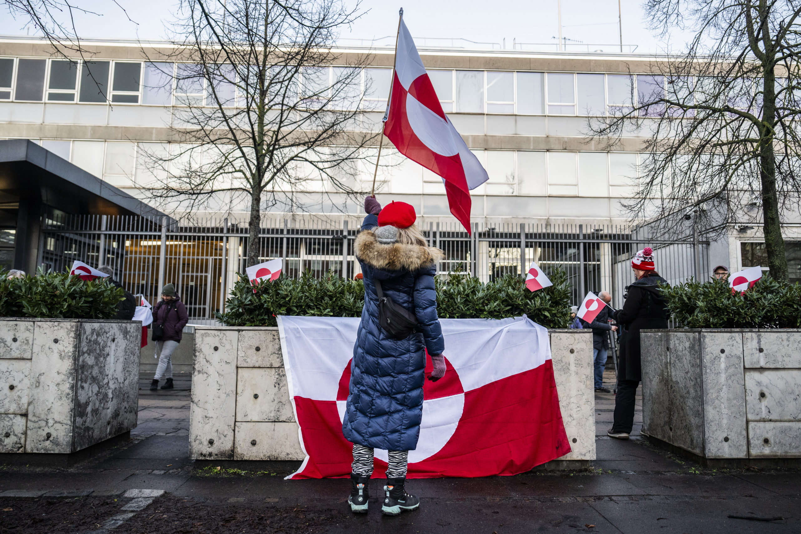 A woman holding the flag of Grenland and wearing a long blue coat with fur trim stands in front of a large Greenland flag hanging on concrete barriers 
