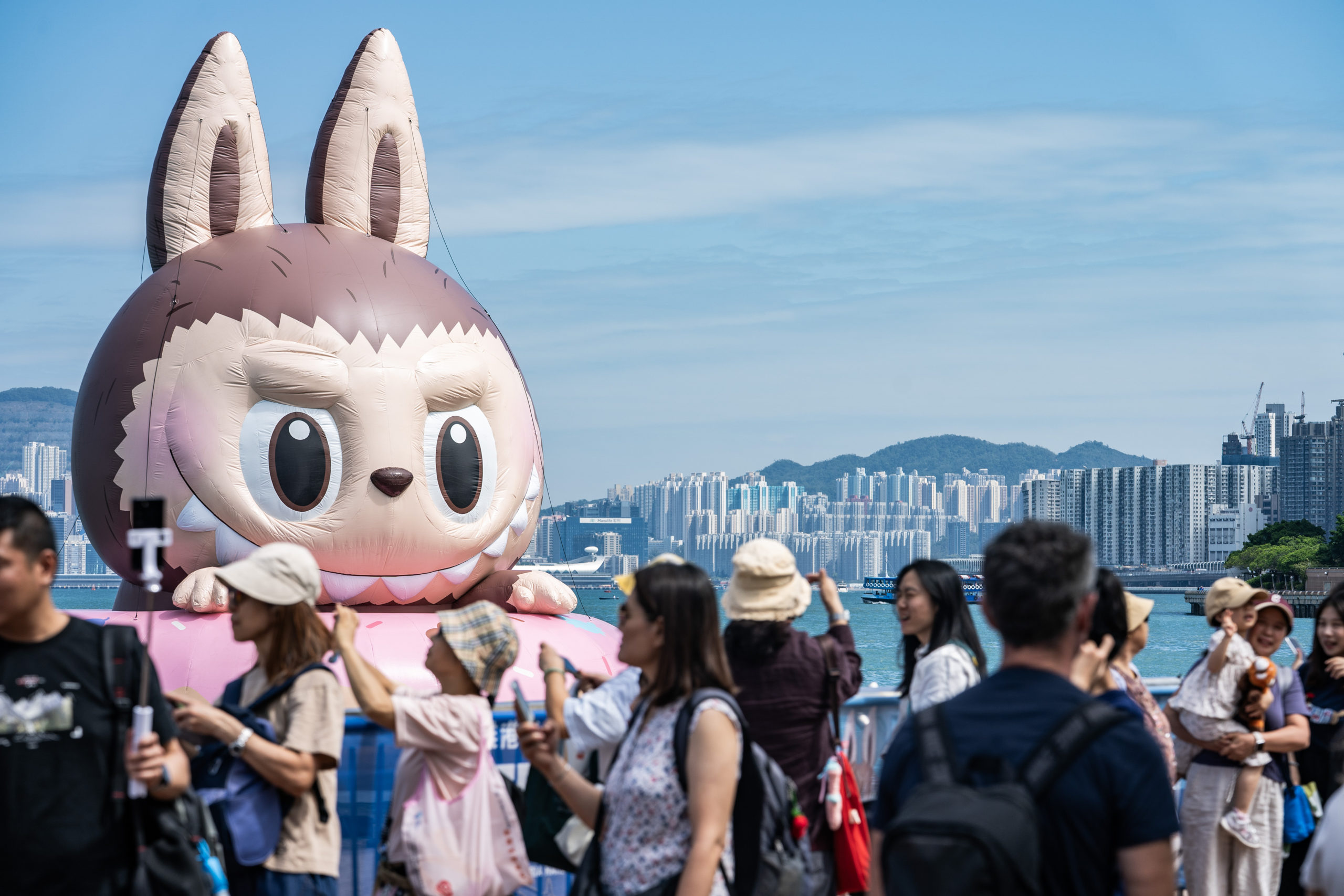 A huge inflatable Labubu in the harbor of Hong Kong, with people milling about in the foreground.