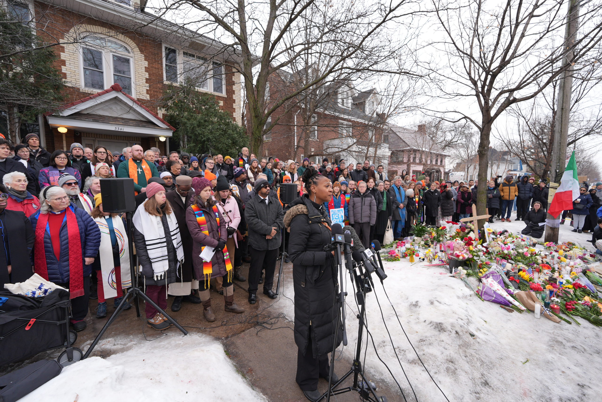 A minister speaks into a microphone at a press conference on a residential street. An array of religious leaders flank her from behind. They are speaking next to a street memorial where Renee Nicole Good was killed.