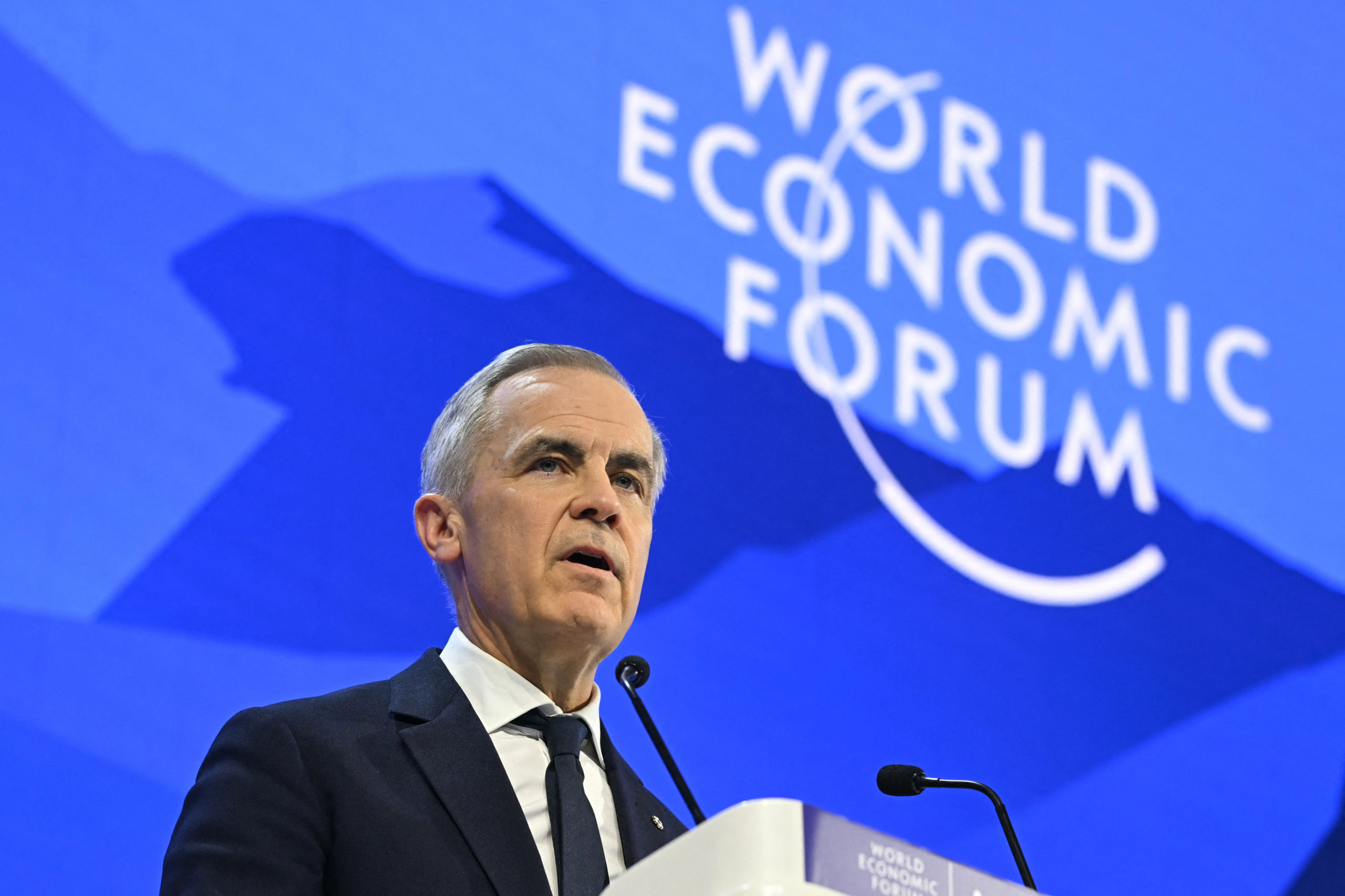 Prime Minister Mark Carney speaks at a microphone with “World Economic Forum” on a blue background behind him.