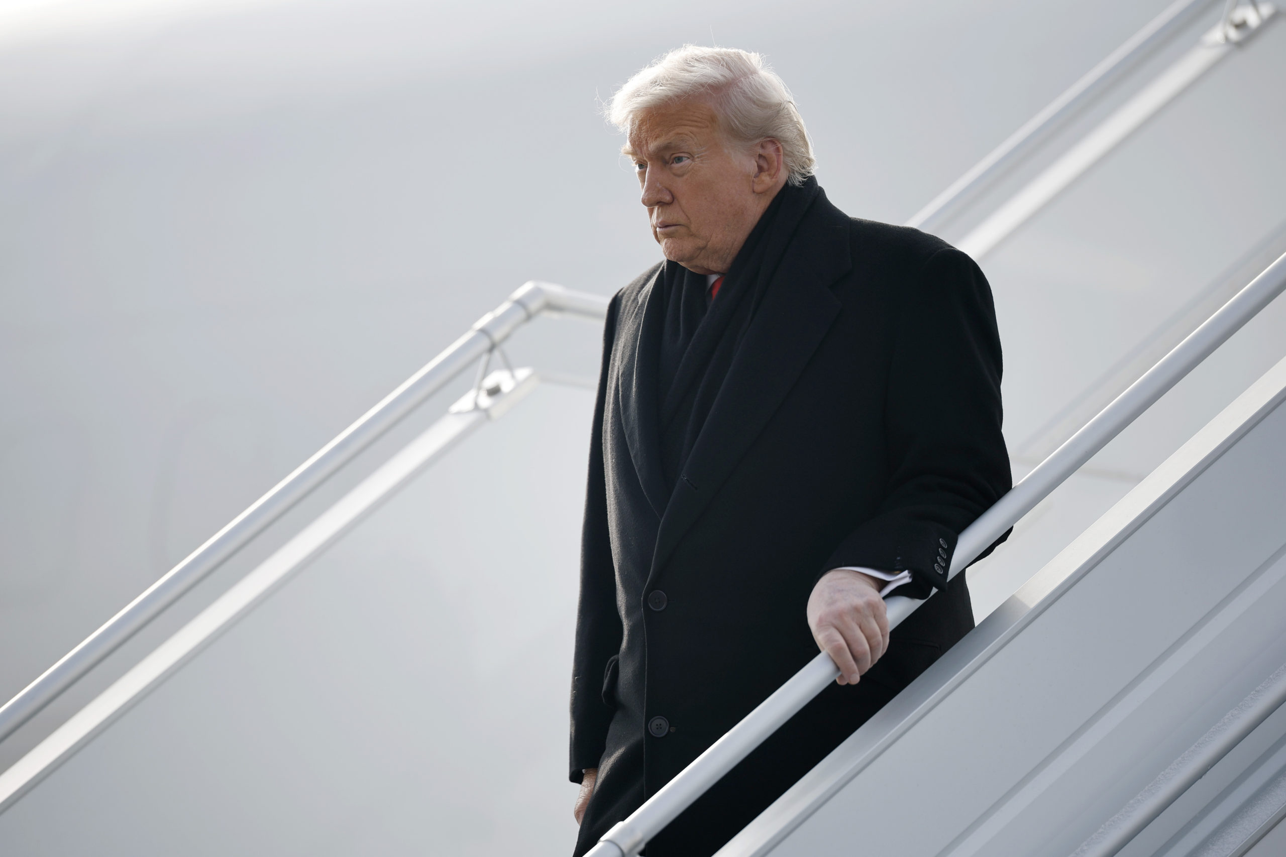 Donald Trump, wearing an overcoat and scarf, holds onto a railing as he descends a set of stairs from Air Force One.