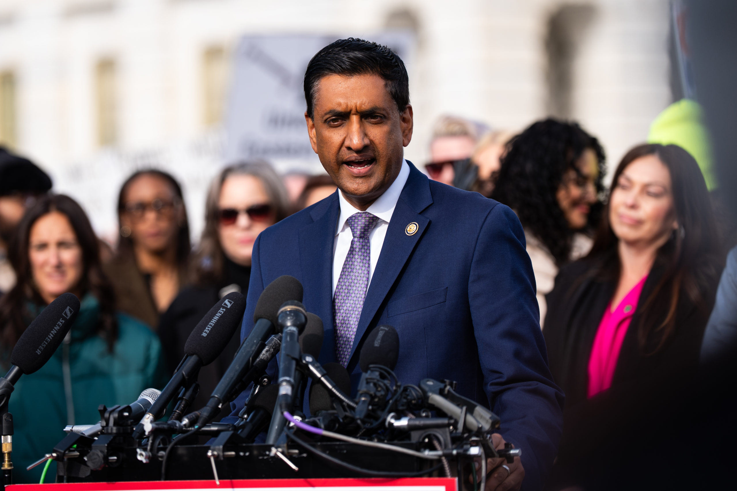 Rep. Ro Khanna, a clean-shaven man wearing a navy suit with a white shirt, speaks from behind a podium covered in microphones; behind him stands an out-of-focus crowd.