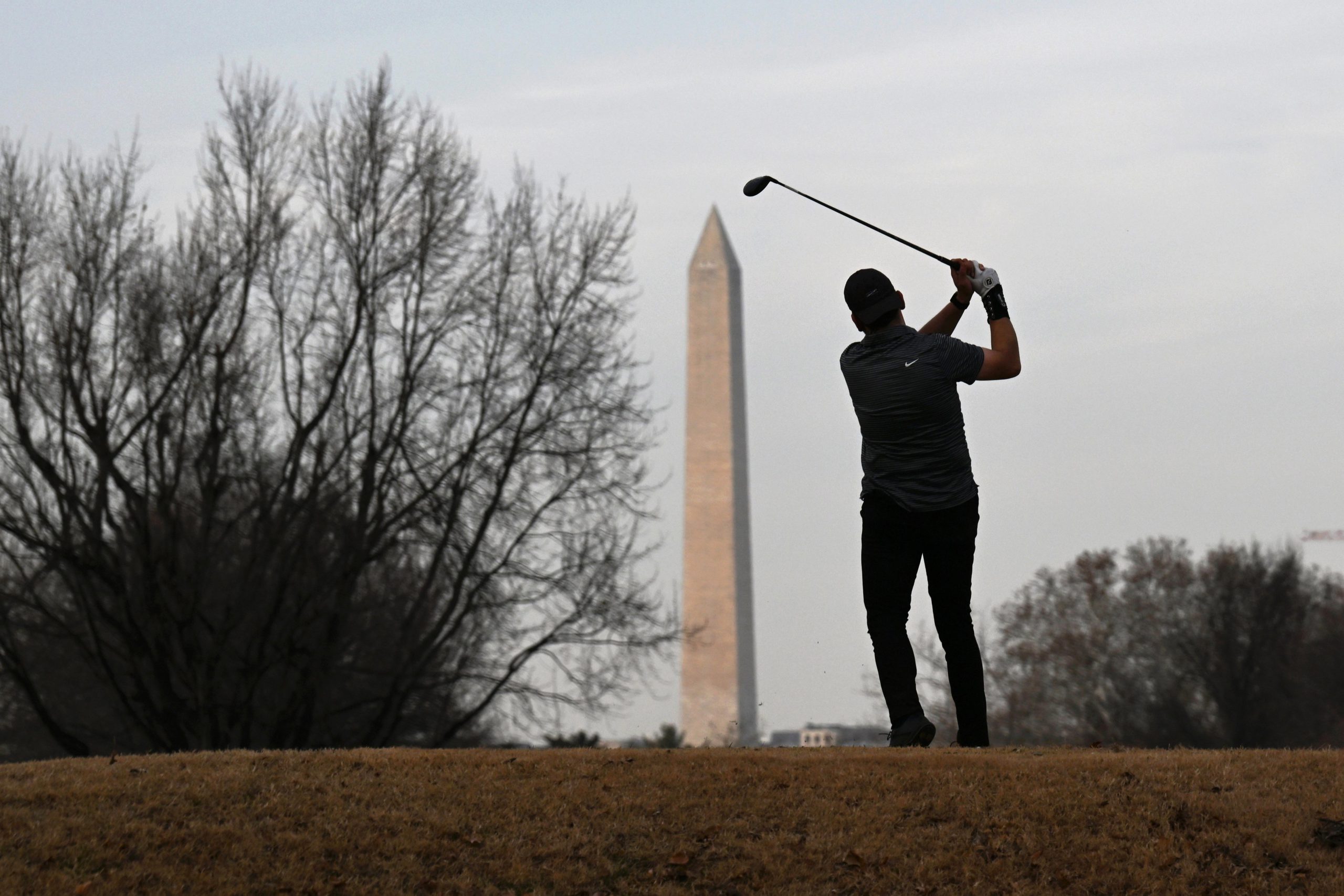 A golfer stands on a rise with his club raised, looking toward the Washington Monument.