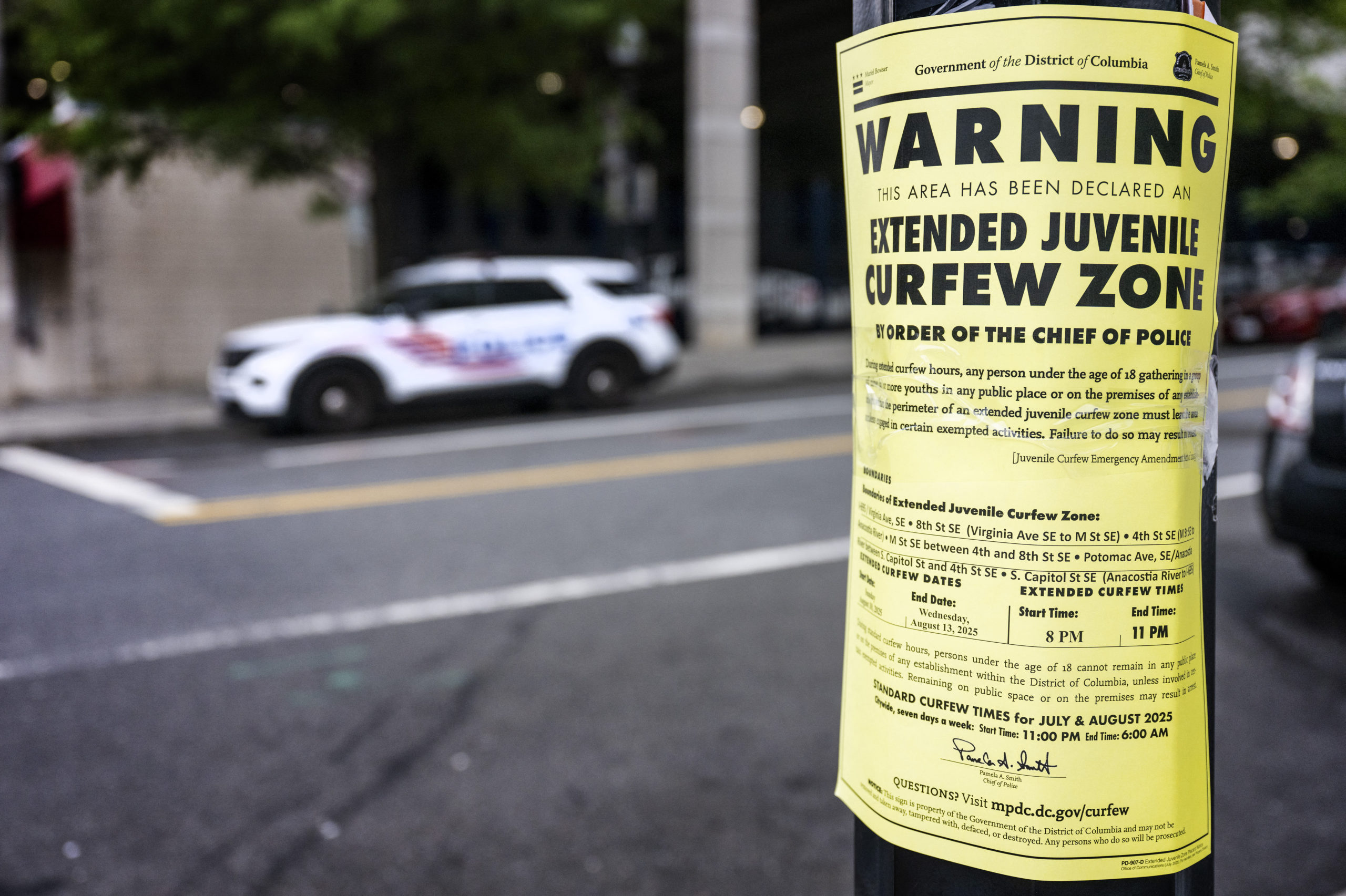 A yellow placard with black text warns that an "Extended Juvenile Curfew" is in effect; in the background, a white police SUV is visible.