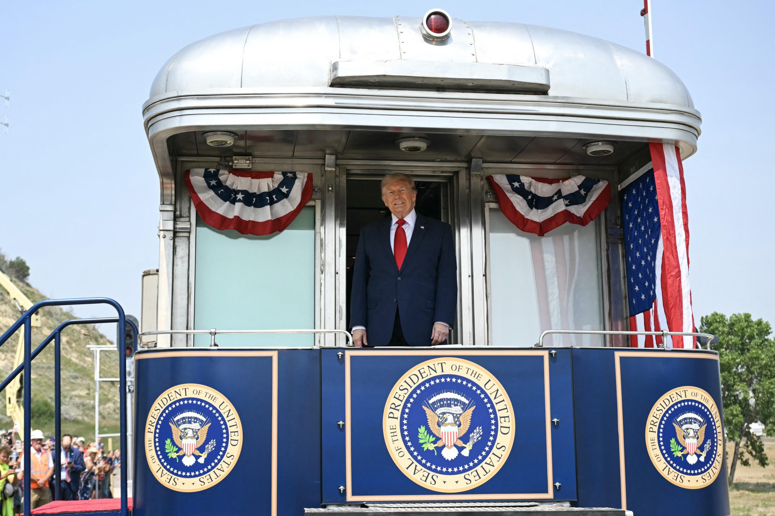 Donald Trump, wearing a suit and tie, stands on the back of a train car decorated with red, white, and blue bunting and the presidential seal.