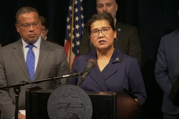 A woman in a navy suit speaks at a podium during a press conference.