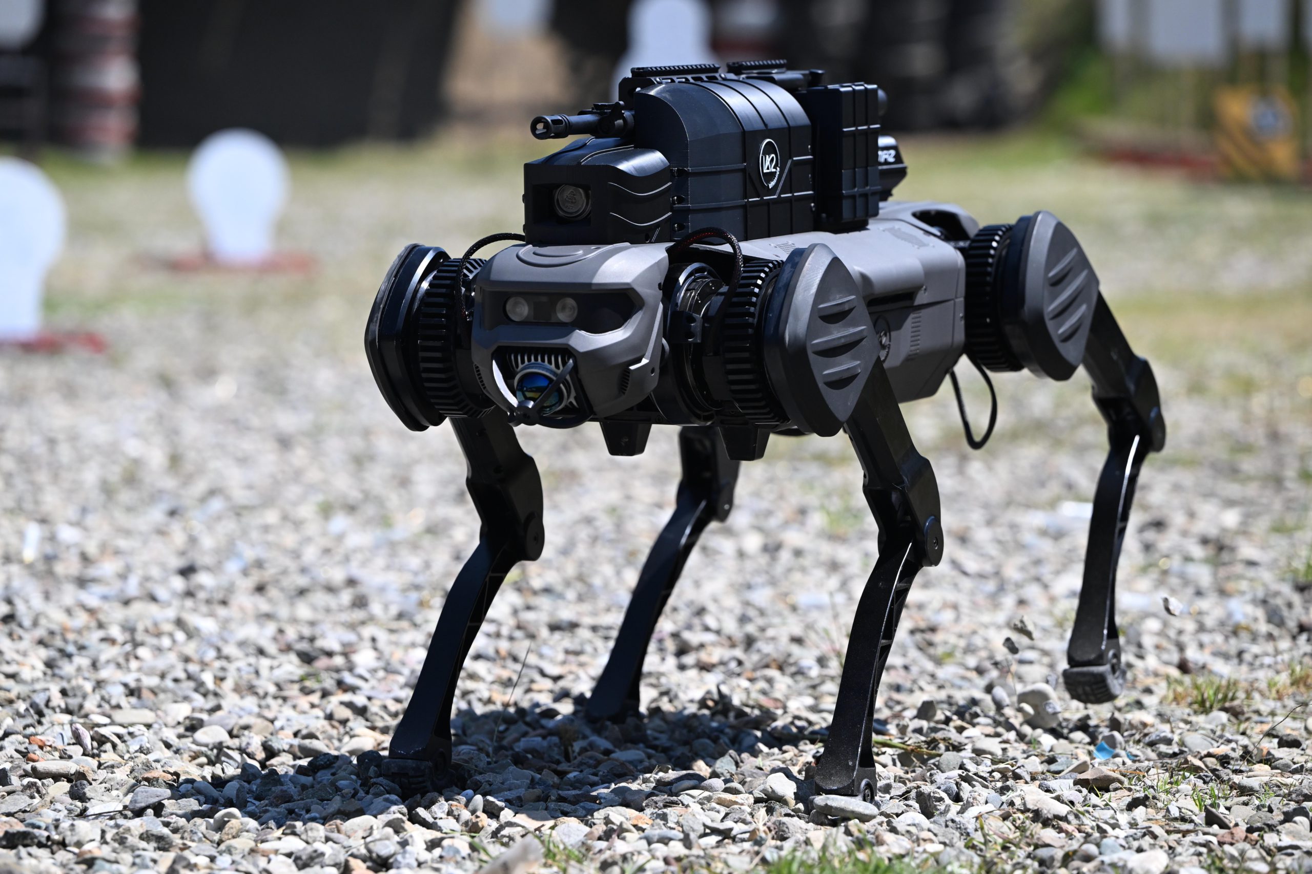 A four-legged robot with armaments on top walks on a gravel surface.