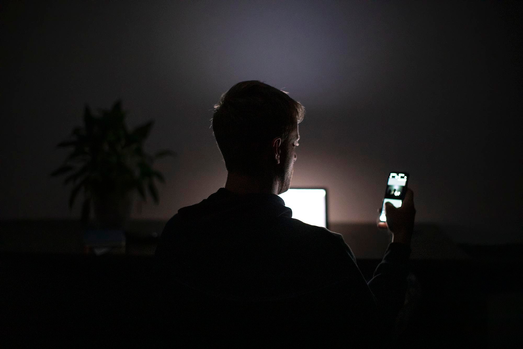 A man in a dark room is starkly backlit by the laptop and iPhone in front of him.