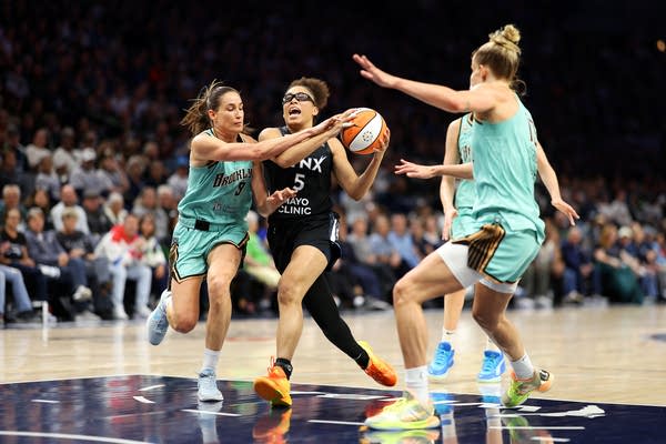 A basketball player in black runs up to the hoop while two defenders in turquoise flank her.
