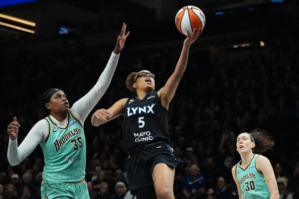 A basketball player in black jumps for a layup while guarded by a player in turquoise.