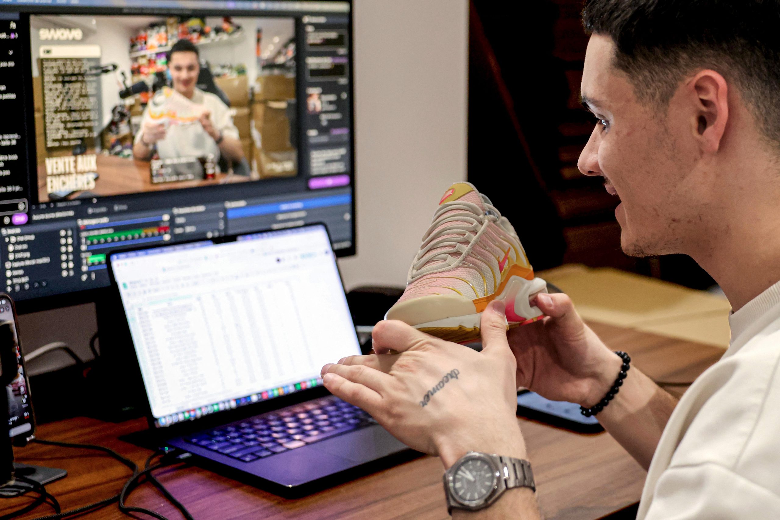A man holds a sneaker in two hands in front of his desk, on top of which are a laptop and a second monitor showing his image on a webcam.