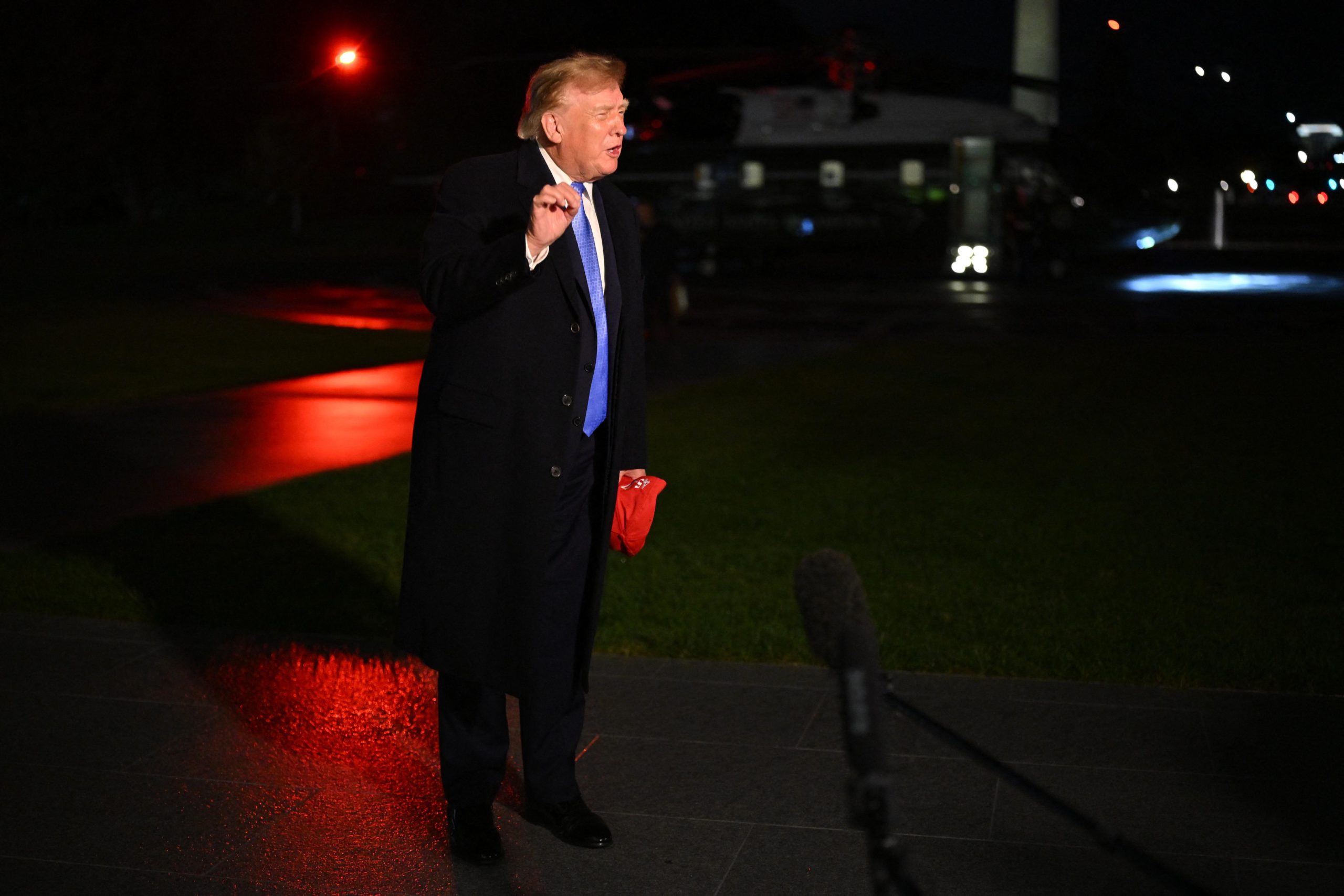 Donald Trump, wearing a dark topcoat over a suit and holding a red hat in one hand, gestures with the other as he speaks in front of the Marine One helicopter at night.