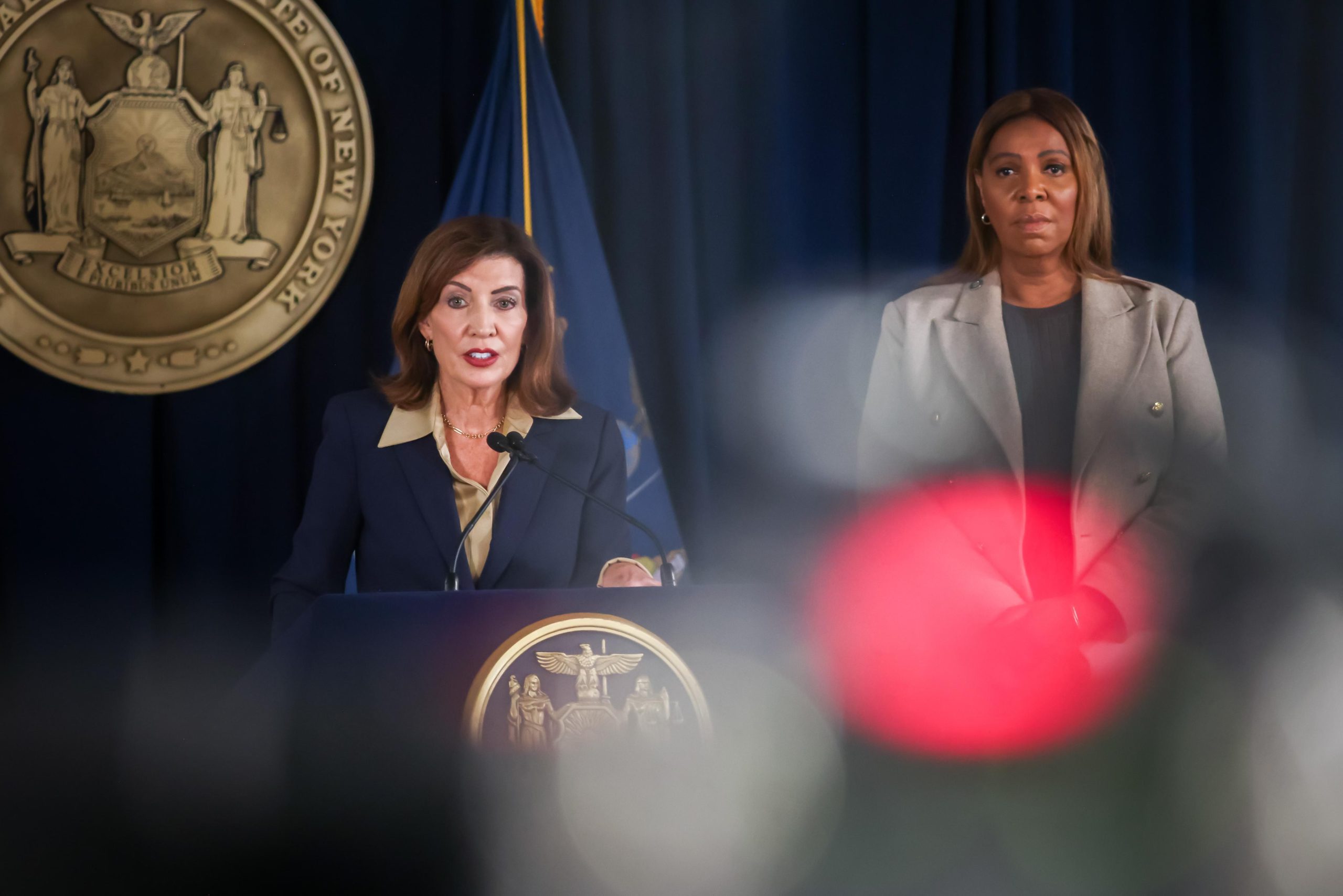 Kathy Hochul, wearing navy, stands behind a podium with the state seal of New York on it; next to her with her hands clasped stands Letitia James.