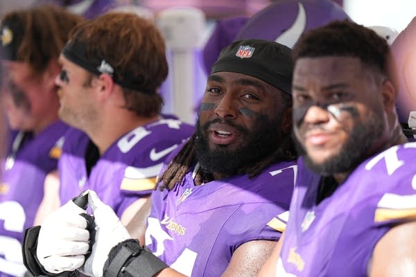 A football player in a purple uniform looks to the side while sitting on a bench with other players.