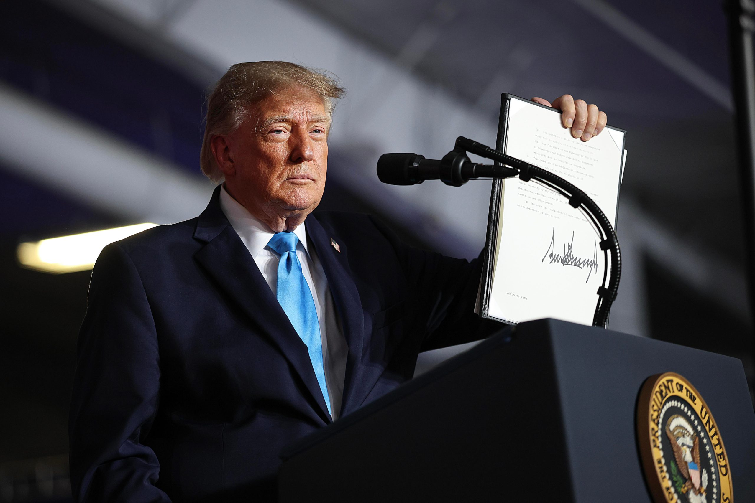 Donald Trump, wearing a suit and tie and standing behind a podium with a microphone on a curving stand, holds up a copy of a signed executive order in his left hand.