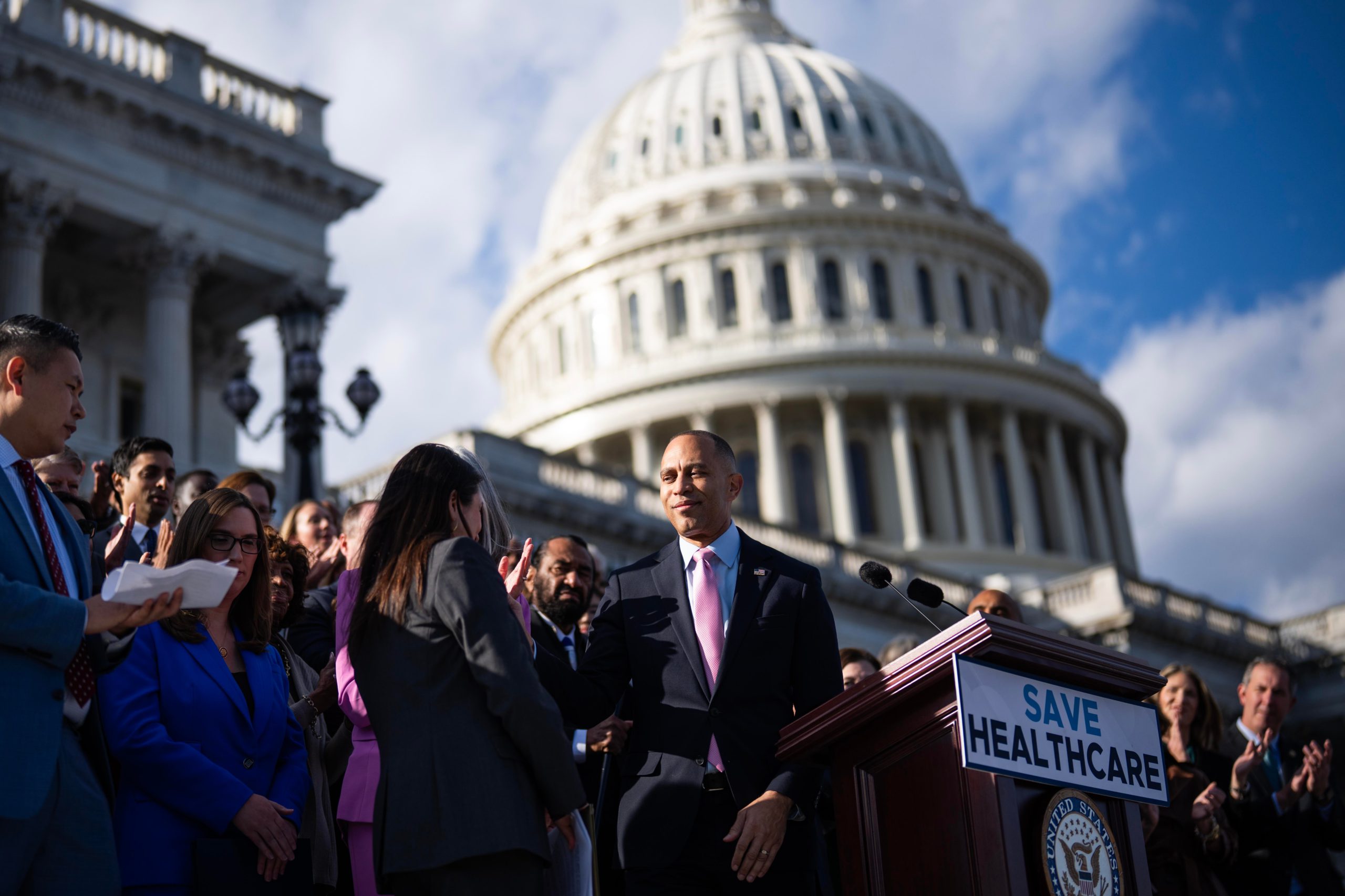 House Democrats gather on the steps of the US Capitol behind a podium with a sign reading “Save Healthcare”; in the background, the Capitol dome is visible against a blue sky streaked with clouds.