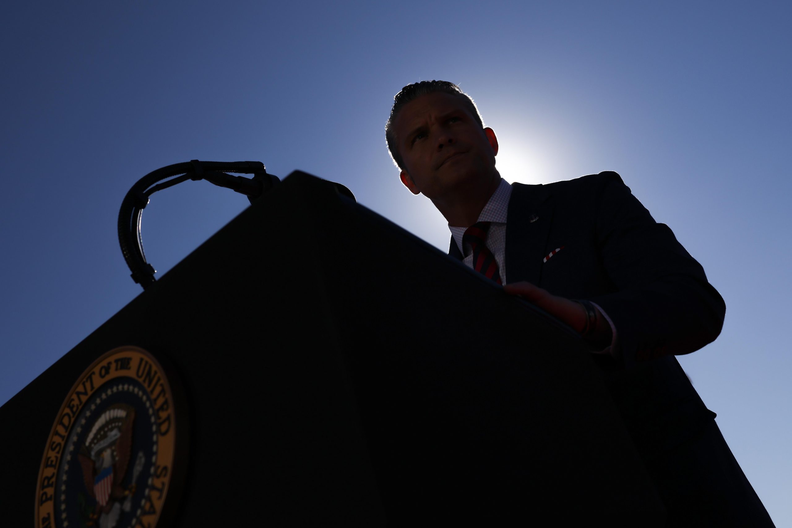 Pete Hegseth, photographed from below and at an angle, is seen in shadow standing behind a podium.
