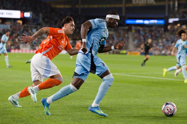 Two soccer players, one in light blue and one in orange, chase after the ball.