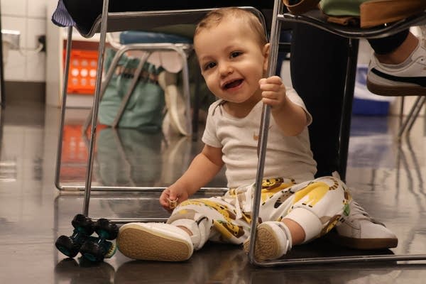 A baby smiles while sitting under a chair.