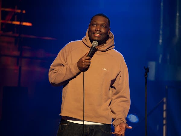 A comedian holds a microphone while performing on a stage.