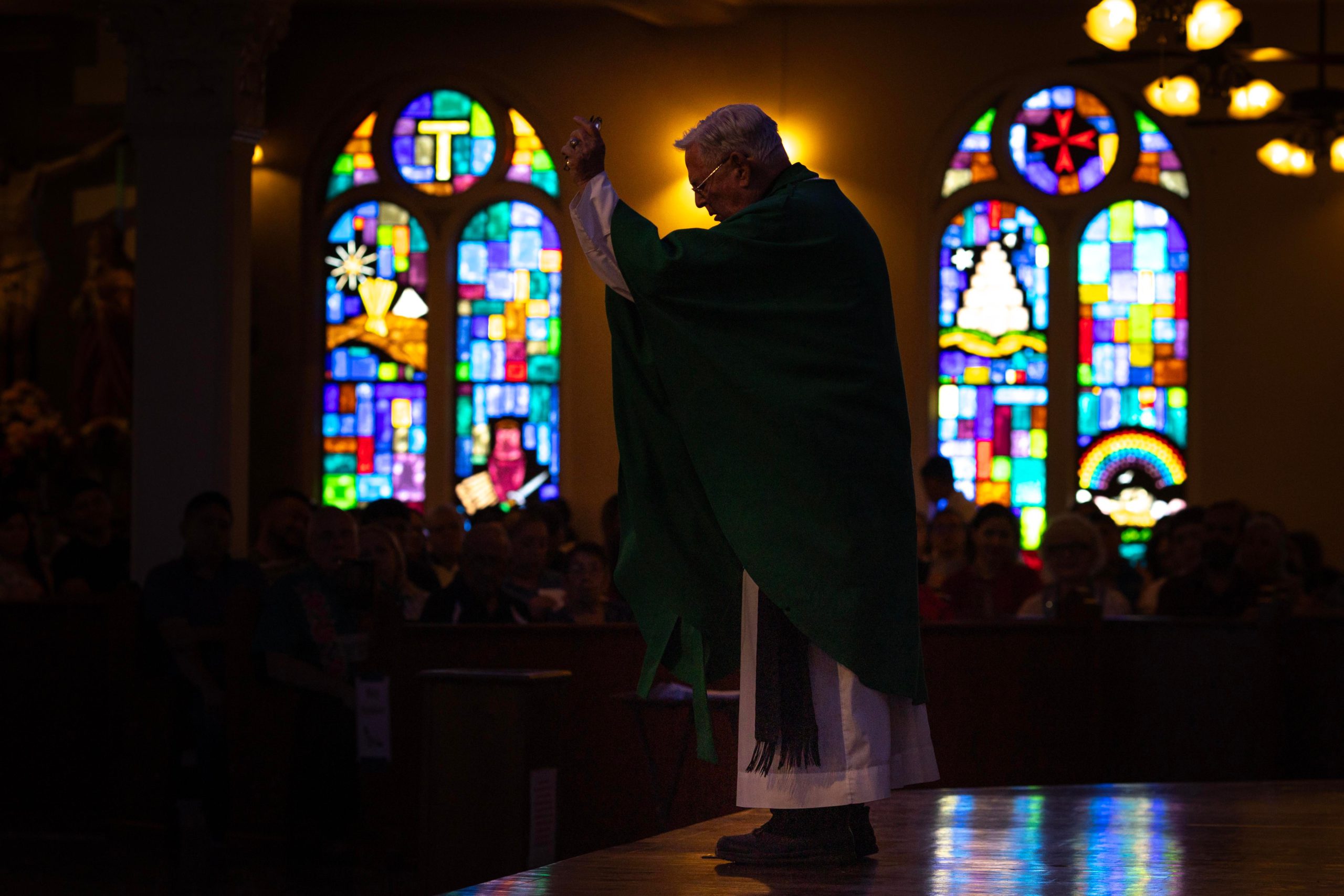 A priest at a pulpit in front of stained glass windows in a church