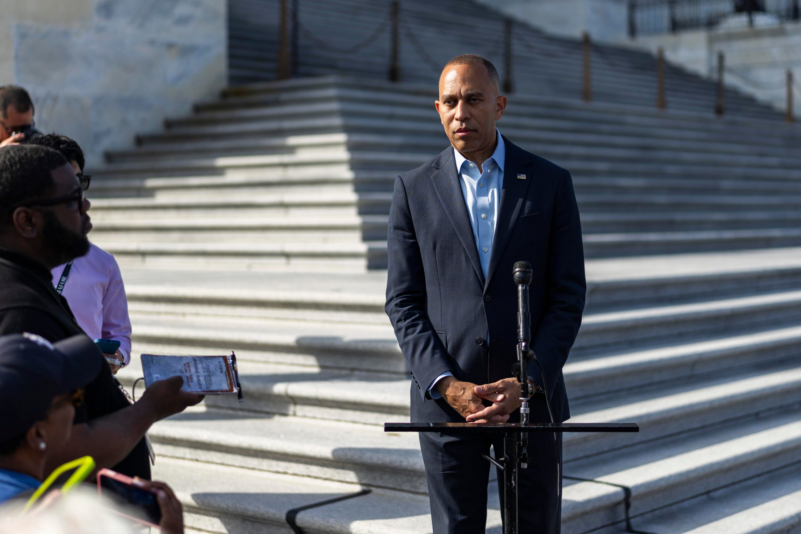 Hakeem Jeffries, wearing a dark suit and a dress shirt without a tie, stands behind a microphone with his hands clasped in front of him; behind is the US Capitol, and journalists stand to his left.
