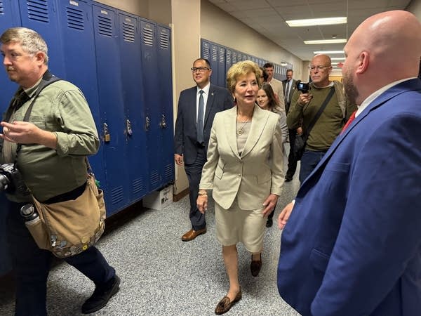 A woman walks through a school hallway by blue lockers while flanked by others in suits.