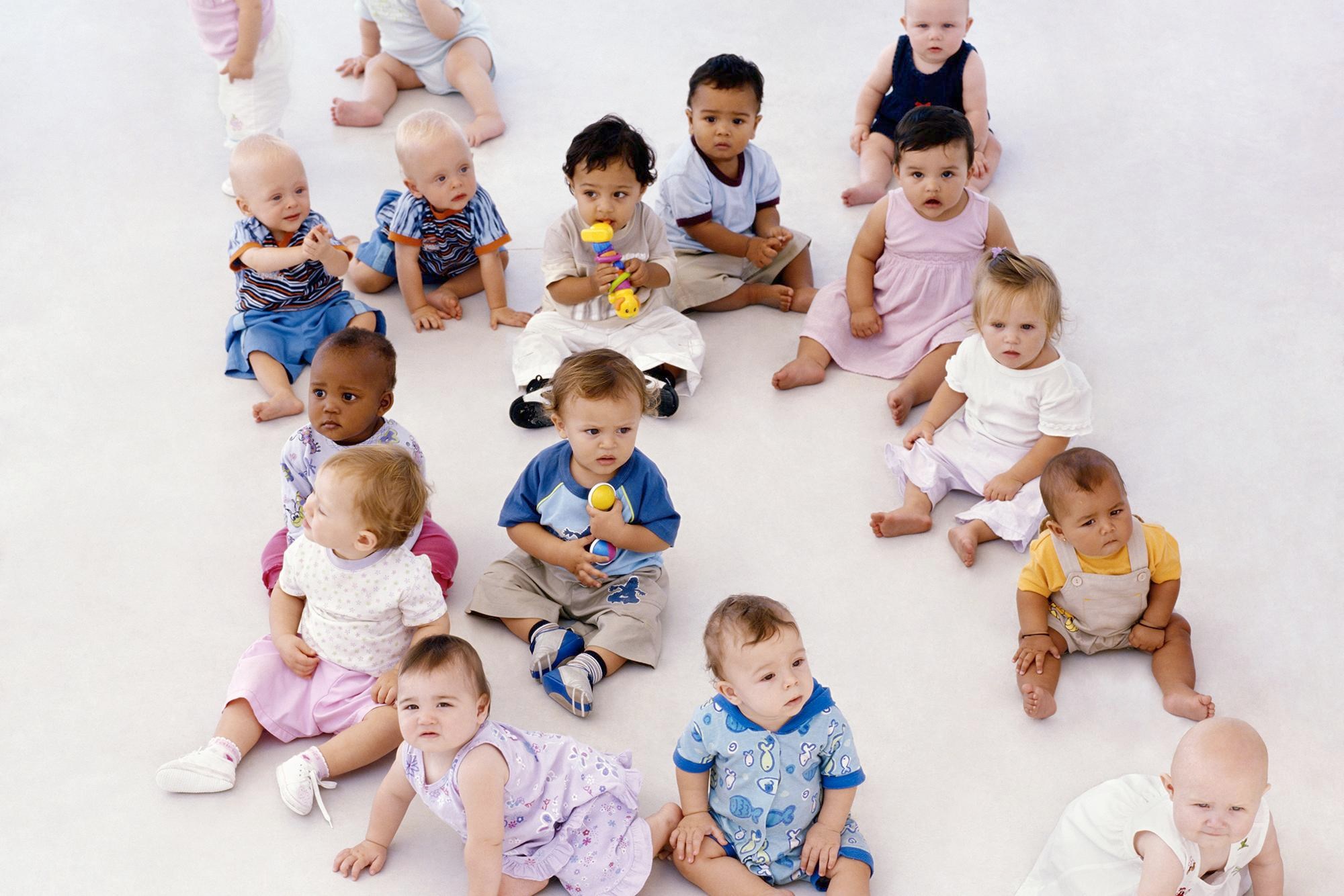 About a dozen babies sit on a seamless white floor.