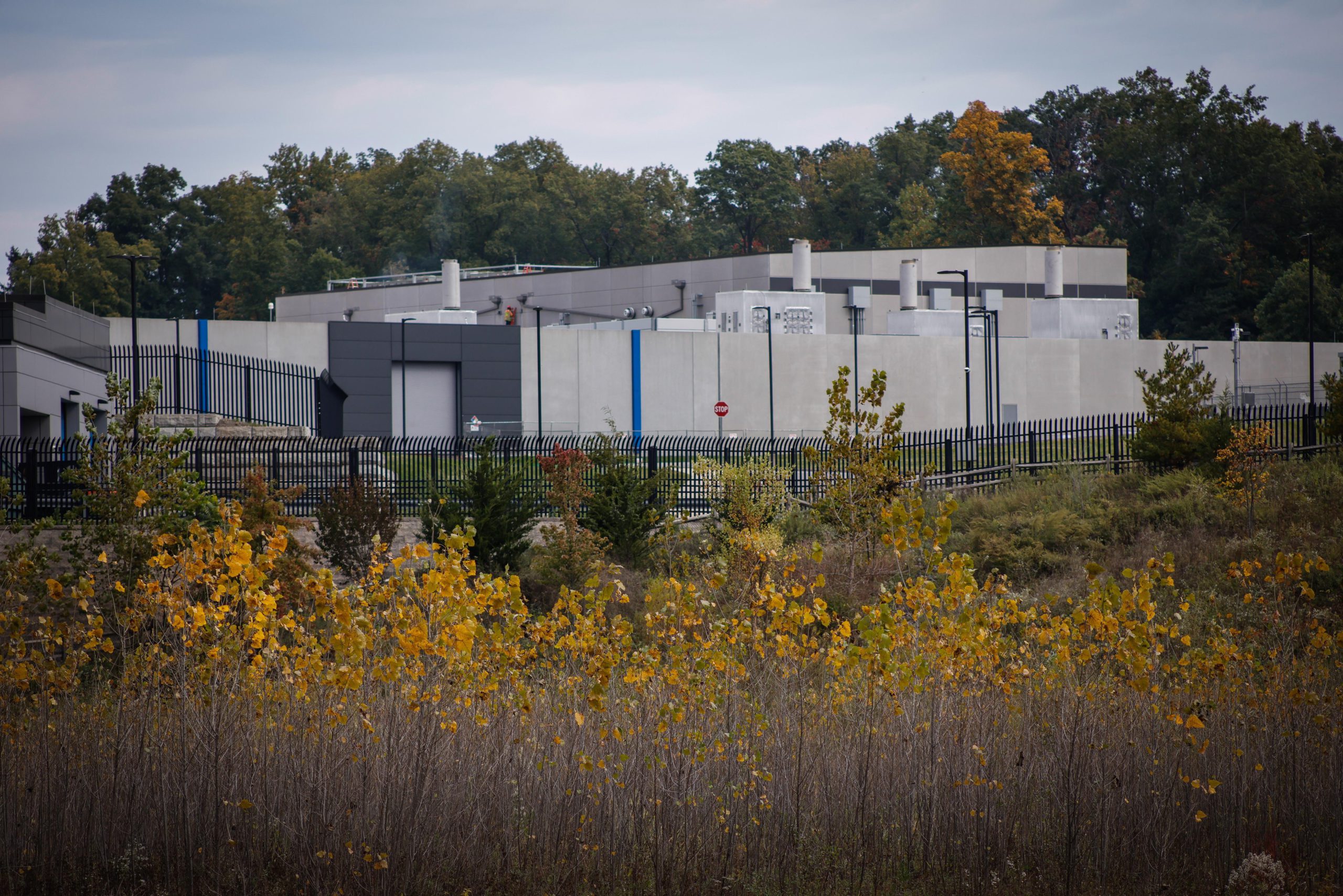 A data center in shades of gray, surrounded by a fence, is seen in front of trees beginning to show autumnal colors; in the foreground is more autumnal greenery.