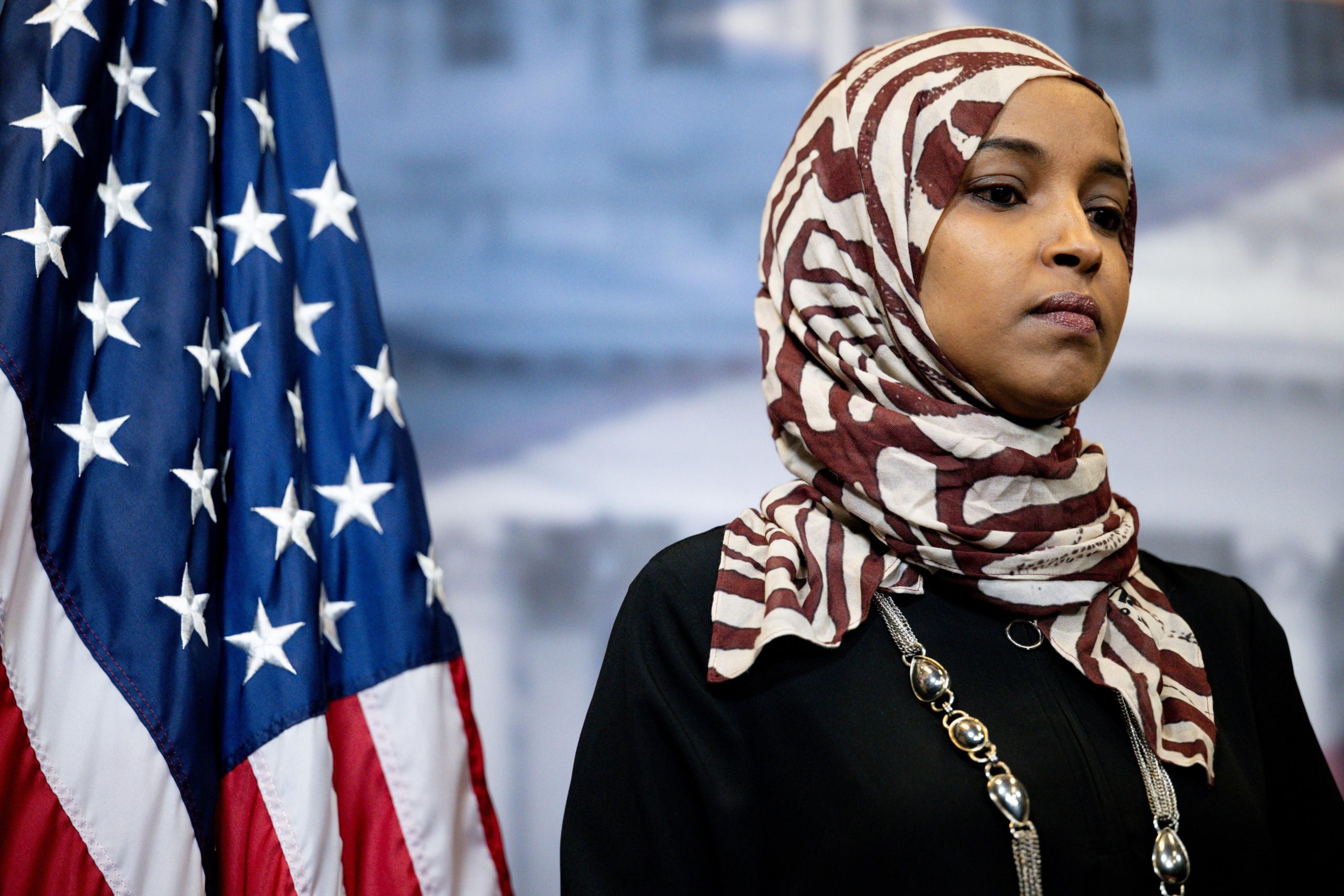 Rep. Ilhan Omar, wearing a white and red head covering and a black top, stands near an American flag at a press conference.