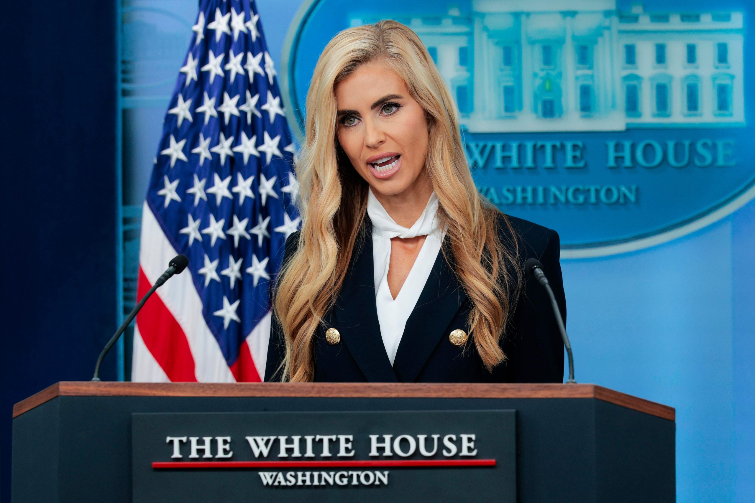 Katie Zacharia, a blonde woman wearing a dark double-breasted jacket over a white blouse, speaks from behind the podium in the White House briefing room.