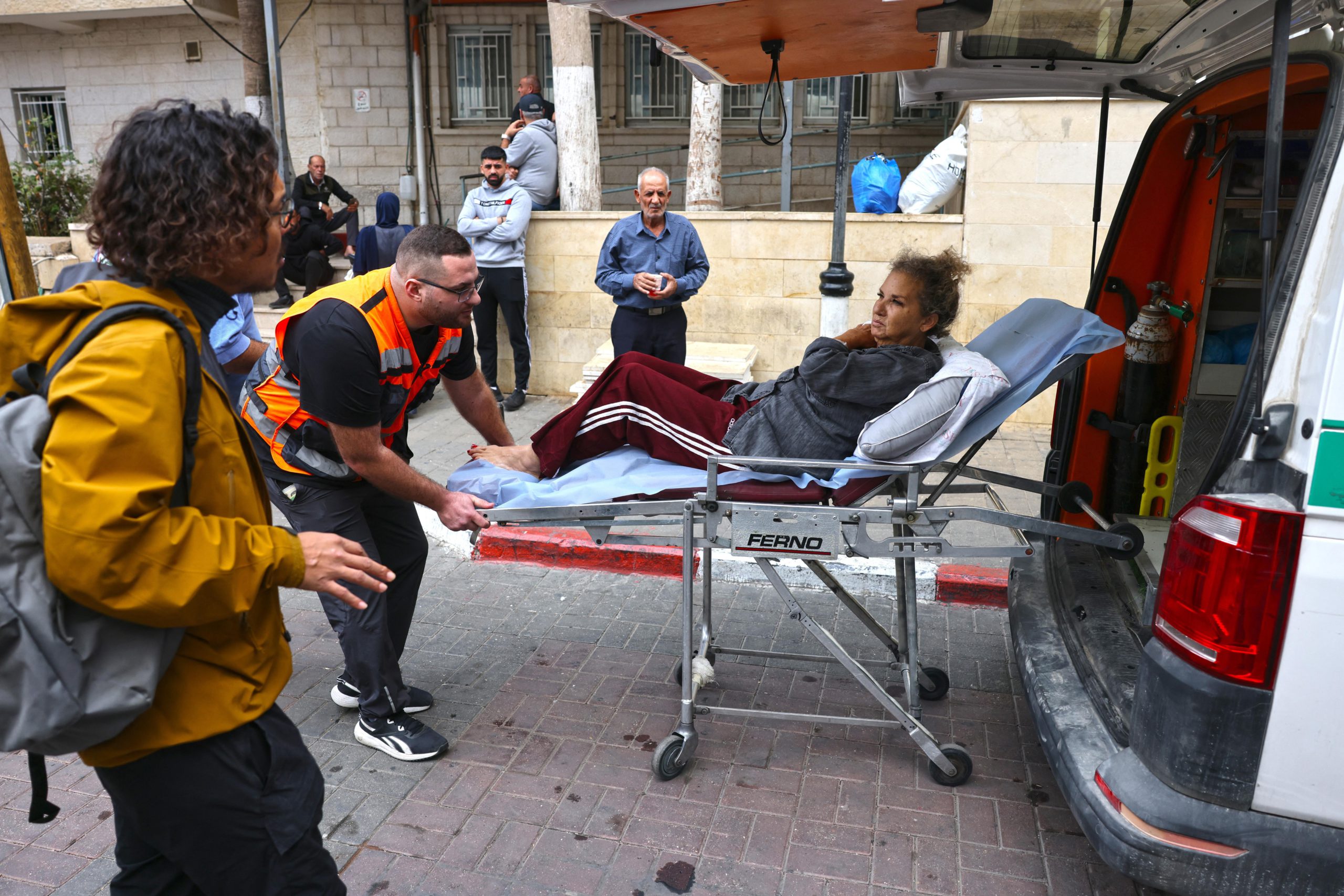 A woman sits on a stretcher as it’s loaded into the back of a vehicle by a man in an orange safety vest.