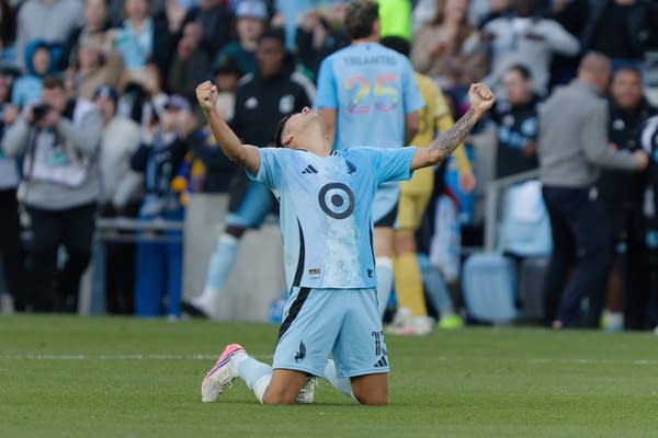 A soccer player in light blue falls to his knees and raises his arms wide in celebration.