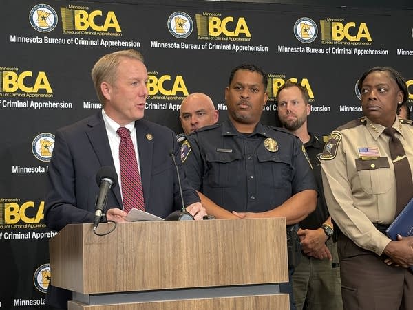 A man in a suit speaks at a podium while flanked by law enforcement officers.