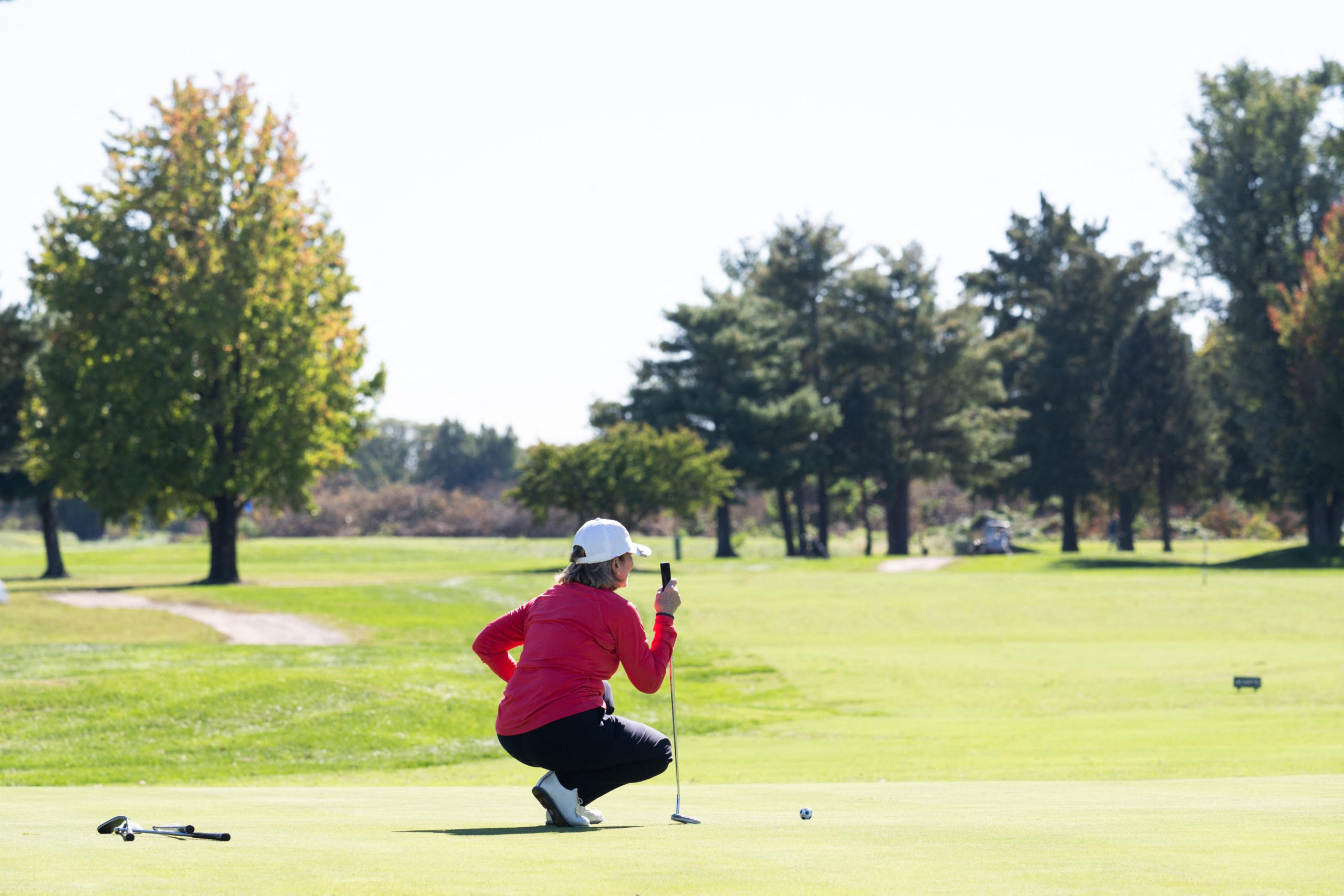 A golfer wearing red and black crouches on a golf course, leaning on a club.