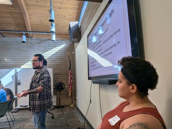 A man gives a presentation to a room of trainees while a woman watches from the side.