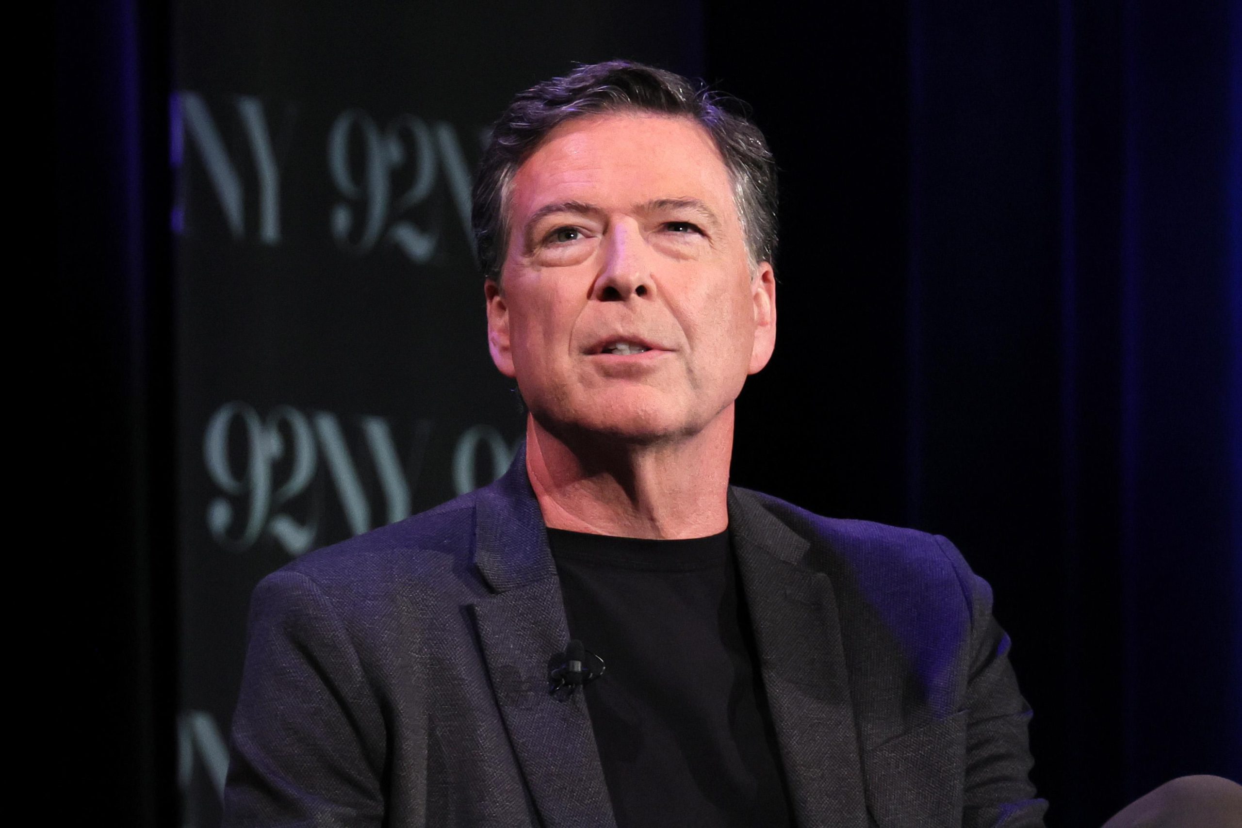 James Comey, wearing a black t-shirt and a dark sportcoat with a lapel mic, speaks onstage at an event.